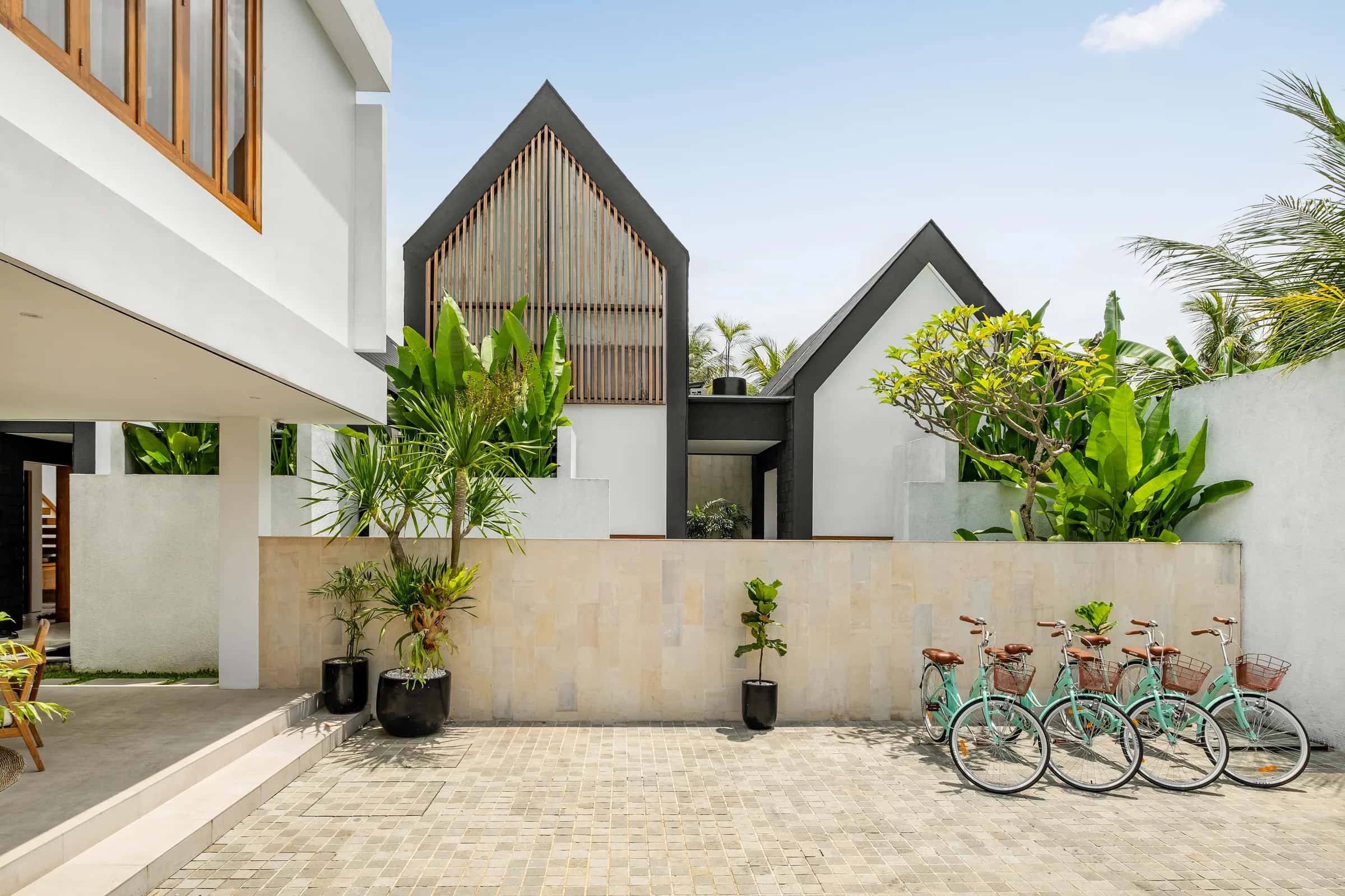 Kedua vila A-frame dari jalan masuk dengan tanaman tropis dan sepeda tamu