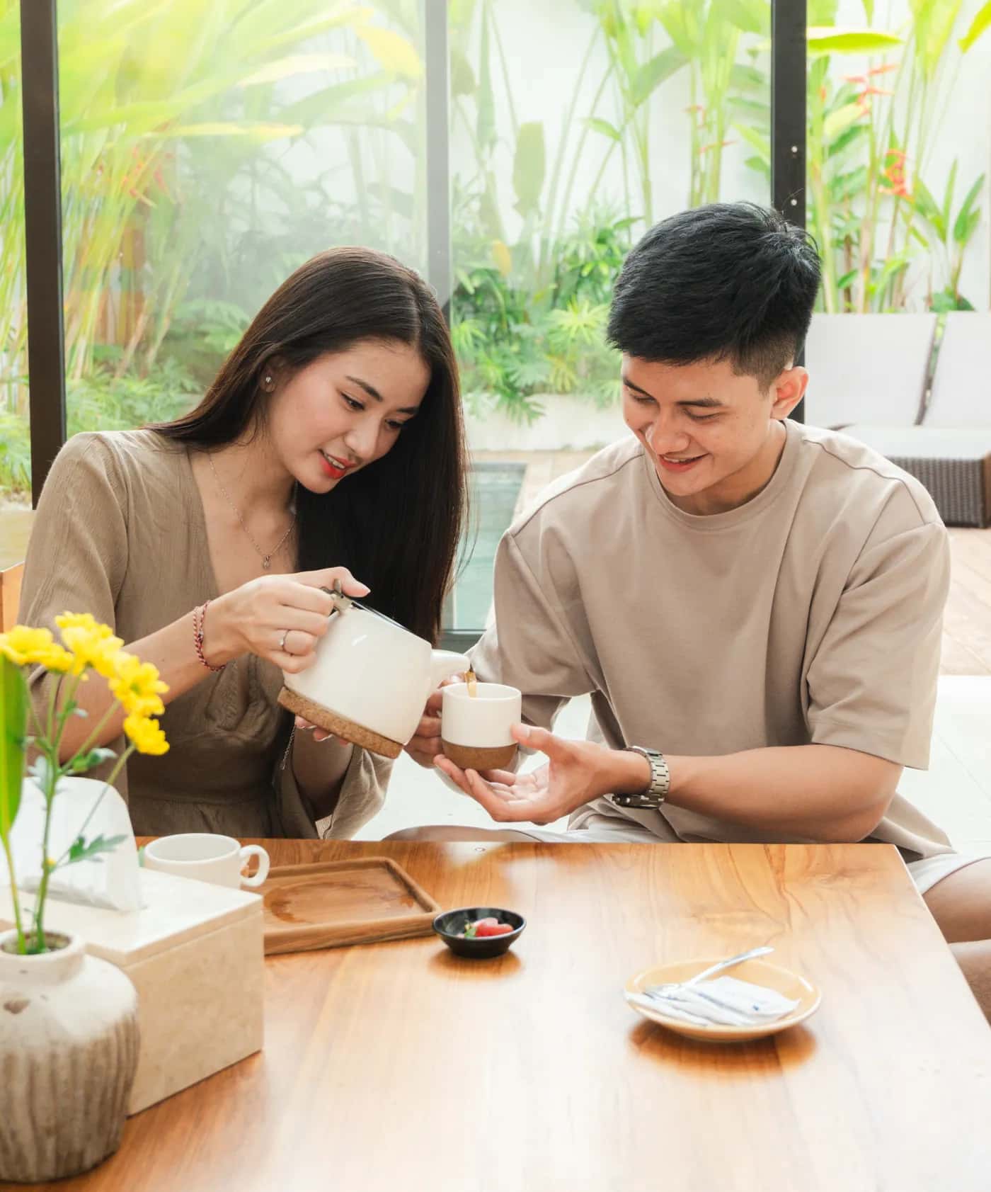 Couple pouring tea at a wooden table at Cafe Teduh, with yellow flowers and a garden beyond