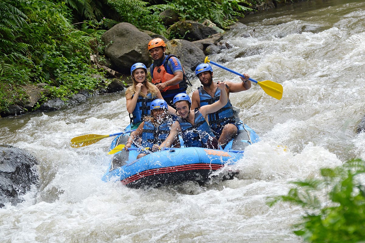 Group of smiling rafters in a blue raft navigating white water rapids through the Ubud jungle