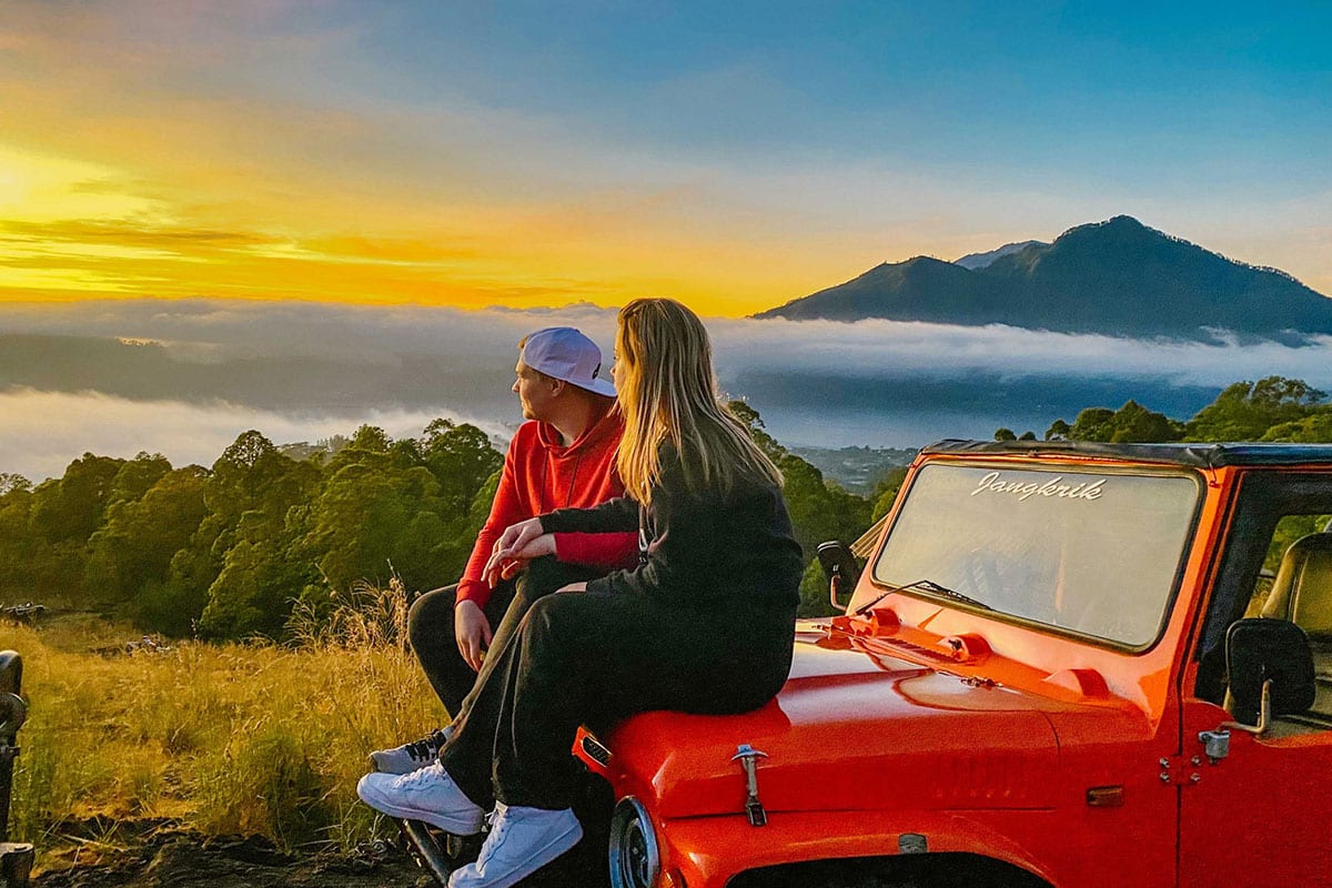 Couple sitting on the hood of a red jeep watching the sunrise over Mount Batur